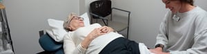 Elderly woman lies on her back on a treatment table while receiving treatment from a physical therapist who is smiling. 
