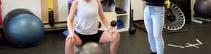 Woman sits on a large bouncy ball while being instructed through an exercise by a pelvic floor physical therapist.