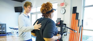 Physical therapist manually cueing a patient's scapular positioning during cable machine exercise for orthopedic shoulder rehabilitation in a bright modern physical therapy gym.
