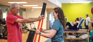 Personal trainer coaching senior male client through resistance band exercise for functional strength training at a physical therapy clinic.