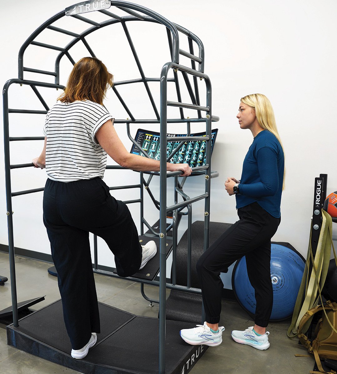 Physical therapist supervising a post-operative patient performing hip flexion exercises on a TRUE Stretch functional training cage for post-op rehabilitation at a physical therapy clinic.