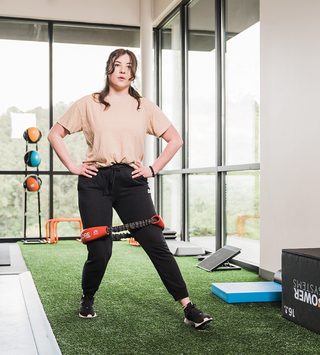 Patient performing resistance band hip abduction exercise for sports injury recovery in a modern physical therapy gym with turf flooring and athletic training equipment.