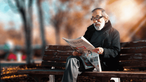 An older man sits on a park bench reading a newspaper.
