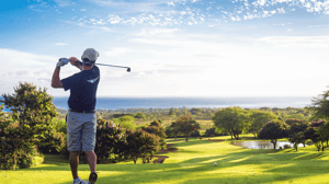 A golfer watches his shot go over a hill on a sunny day.