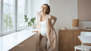 A pregnant woman leans on a windowsill gripping her back in pain.