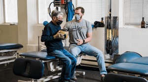 A man sits holding a ball while his therapist guides him.