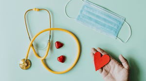 Flatlay of a stethoscope, surgical mask, and hand holding a paper heart