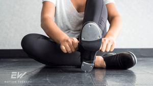 A female tap dancer sits on the floor putting on her tap shoes.