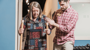 A man guides a woman walking with mobility sticks. 