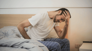 A man sits on the side of his bed holding his head in his hand.