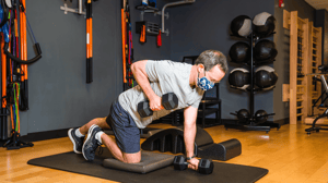 A man kneels on a mat and lifts weights.
