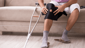 A man sits on a couch to put on a knee brace with a crutch propped up beside him.