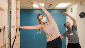 A woman stretches her left arm over her head while a man mirrors her in the background.