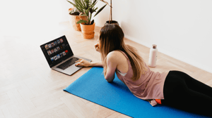 A woman lies on her stomach on a yoga mat looking at a laptop.