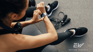 A woman sits on the ground at the gym, looking at her smartwatch.