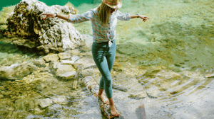 A woman balances on a log while she walks barefoot in a stream.