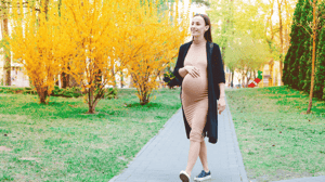 A pregnant woman walks outside in a park, surrounded by yellow-leafed trees.