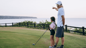 An adult watches a young girl hold a golf club.