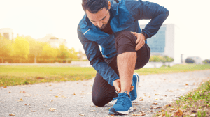 A man on a run kneels to inspect his left ankle.