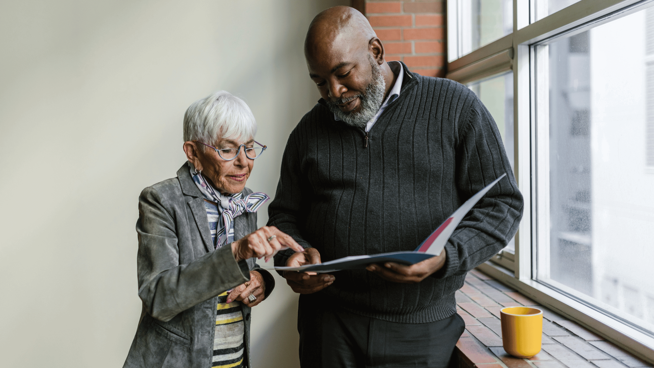 An older woman and a Black man look at a book standing by a window.