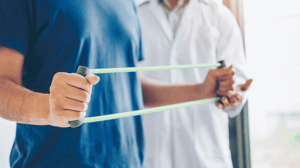 A man in a blue shirt stretches a resistance band while a physical therapist observes.