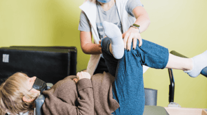 A woman lays on a table while a physical therapist touches her knee. 