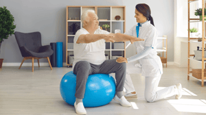 A nurse helps an older man balance on an exercise ball.