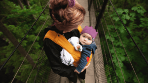 A woman holds her baby on a bridge in the woods.