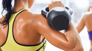 From behind, a woman in a yellow sports bra holds a kettlebell over her shoulder.