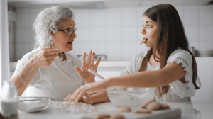 A grandmother sits in the kitchen eating cookies with her granddaughter.