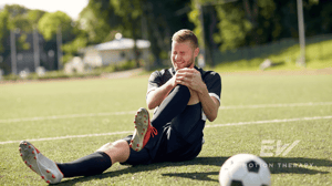 A soccer player sitting on the field grabs his knee in pain.