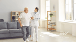 An older man walks through his living room with the help of an aid.