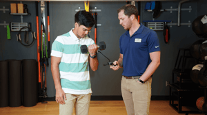 A man lifts a weight while another man monitors a blood pressure cuff on his arm.
