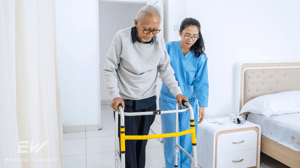 An elderly man uses a walker, accompanied by a female nurse.