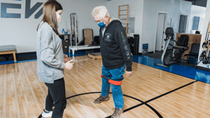 A woman guides an older man through a balance exercise.