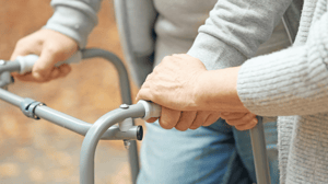 A pair of hands hold a walker, with one supportive hand holding on.