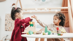 Two little girls sit at a table together and play with blocks.
