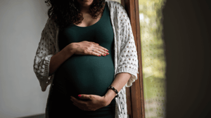 A pregnant woman stands by a window holding her belly.