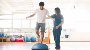 A man steps onto a Bosu ball with the help of an aid.