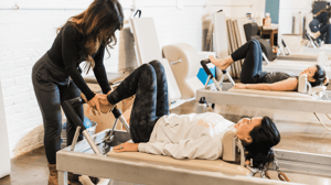 A woman holds another woman's feet on a Pilates reformer.