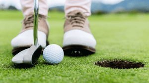 Close-up of a man's feet in golf shoes, about to hit a ball into a hole.