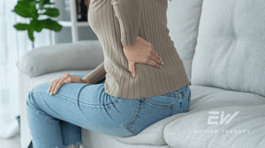 A woman sits on a couch holding her back in pain.