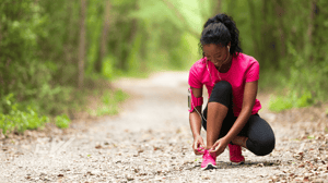 A runner kneels down to tie her shoe on a trail.