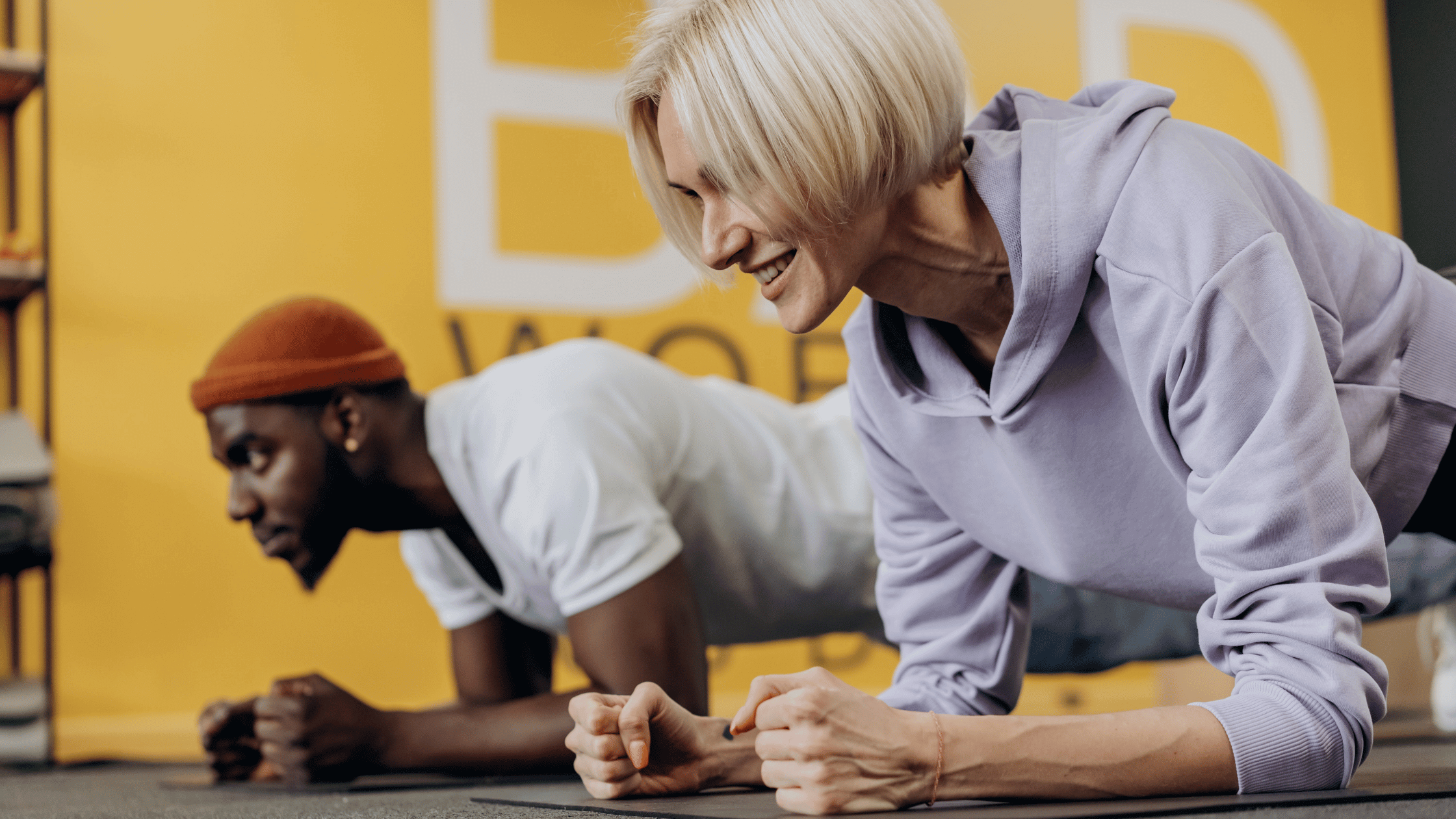 A woman and man plank on the ground in a gym.