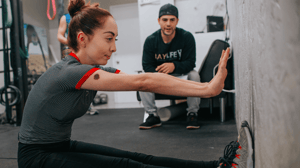 A woman stretches against a wall while her male trainer watches.