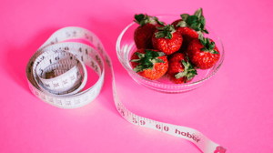 A bowl of strawberries next to a tape measure on a pink background.