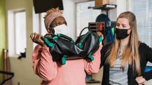 A woman holds up a weighted bag while her therapist watches.