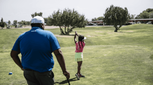From behind, a young girl swings a golf club while a male coach watches.