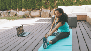 A woman stretches outside on a yoga mat, with her computer set up in front of her.